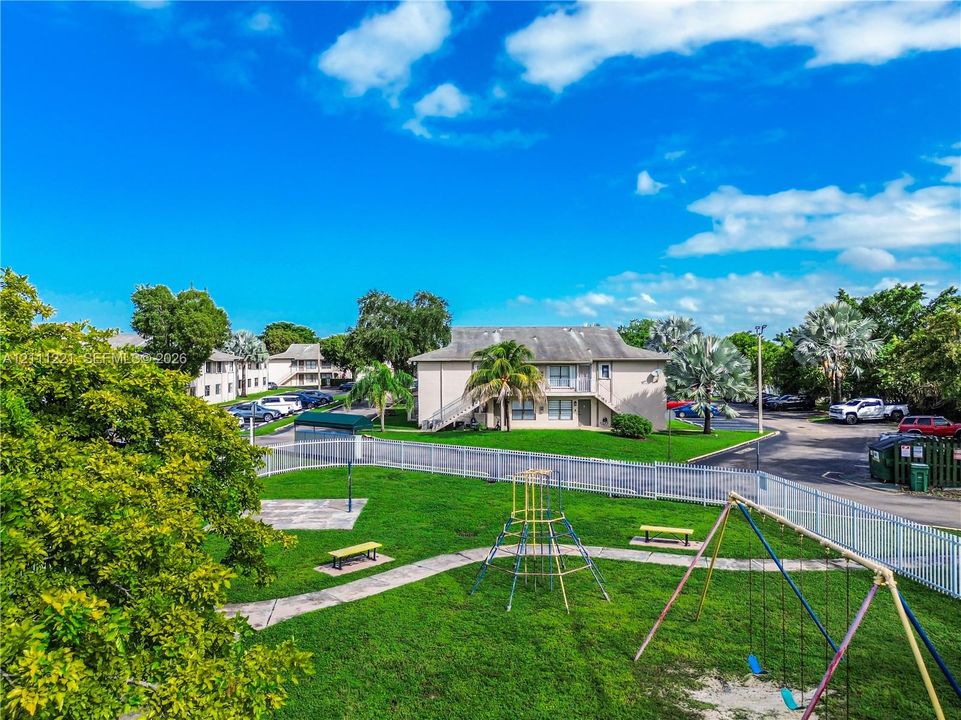 Common area featuring a playground with a climbing frame, swing set, and concrete walkways, enclosed by white metal fencing and landscaped grounds