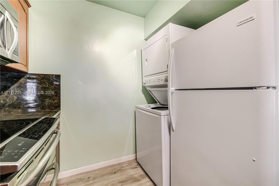 Stacked laundry unit, full-size refrigerator, electric range with dark stone-finish backsplash, and wood-finish flooring