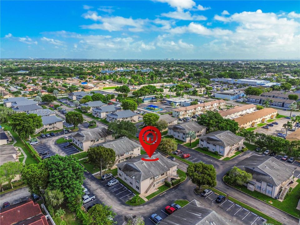 Aerial perspective of a multi-unit residential building featuring a multi-pitched roof, paved parking area, and surrounding mature landscaping