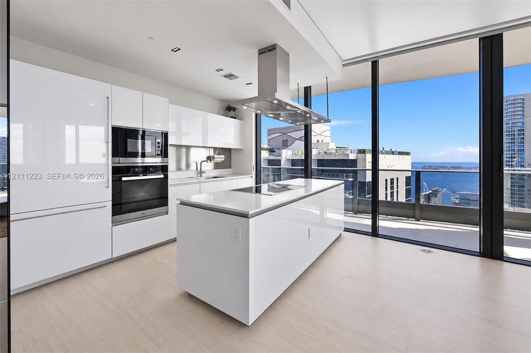 Kitchen featuring high-gloss cabinetry, stone-surfaced center island, stainless steel built-in appliances, stainless steel range hood, and floor-to-ceiling glass paneling
