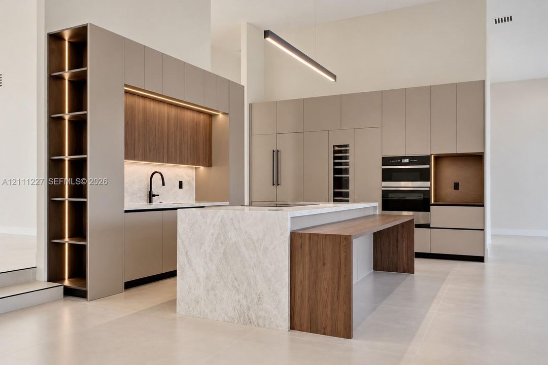 Modern kitchen featuring flat-panel cabinetry, a waterfall-edge stone island with an integrated wood-finish breakfast bar, built-in double ovens, and recessed LED accent lighting