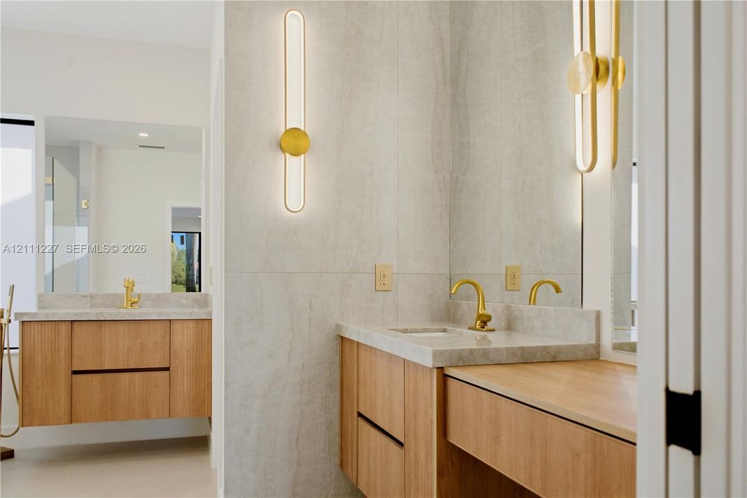 Floating wood-finish vanities with light stone countertops, integrated sinks, brushed gold faucets, and elongated vertical LED wall sconces