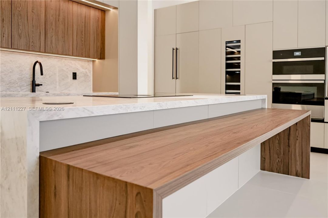 Kitchen featuring a waterfall-edge island with wood-finish counter seating, integrated stainless steel appliances, full-height cabinetry, stone-finish backsplash, and matte black faucet