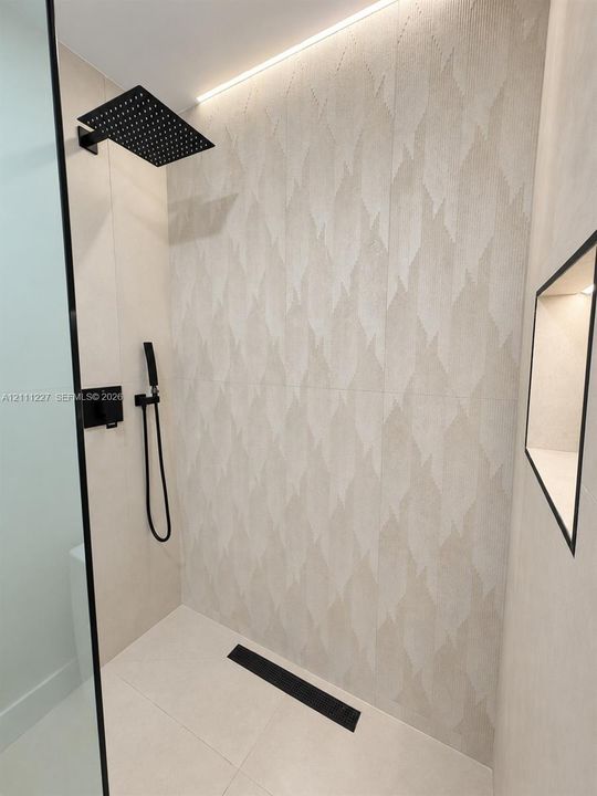 Walk-in shower featuring patterned large-format wall tile, matte black rainfall and handheld showerheads, integrated recessed niche lighting, and a linear floor drain