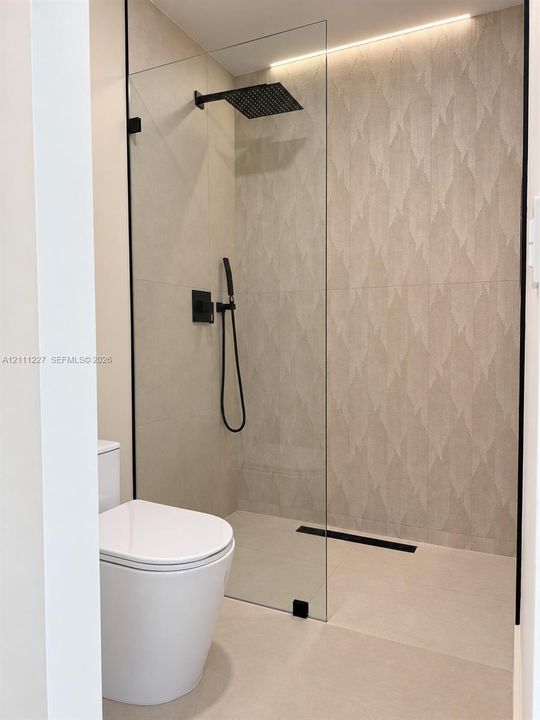 Walk-in shower featuring a frameless glass enclosure, patterned tile accent wall, matte black rainfall showerhead, integrated handheld sprayer, and linear floor drain, accompanied by a modern dual-flush toilet