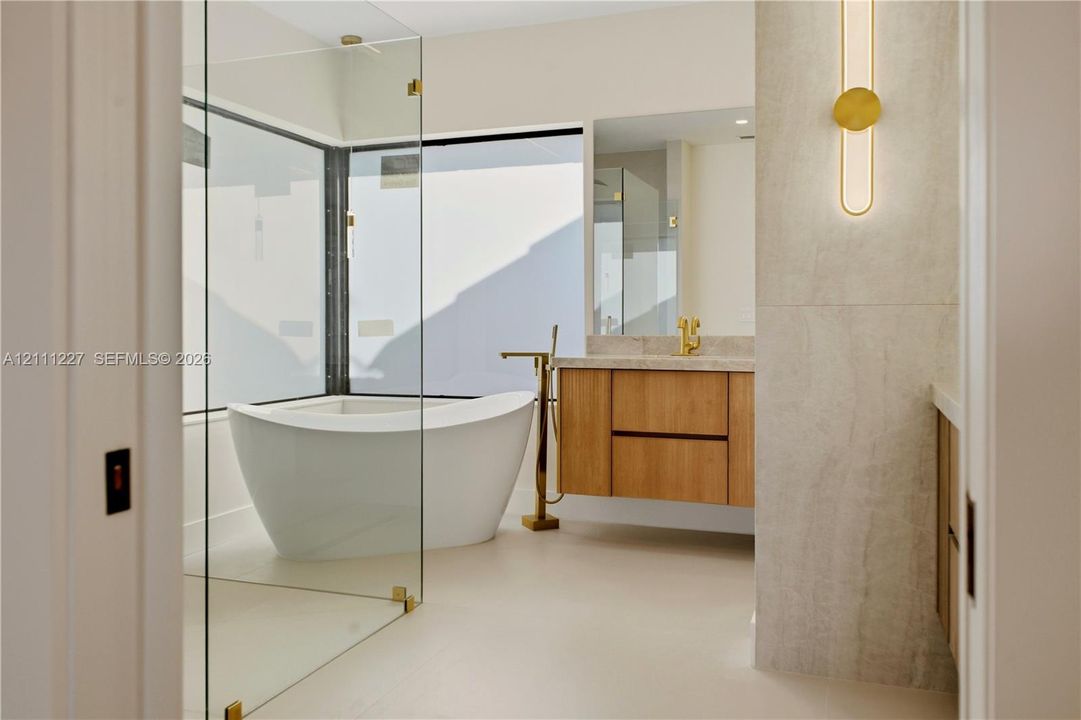 Freestanding soaking tub, frameless glass-enclosed shower, wall-mounted wood-finish vanity, stone countertop, and brass-tone plumbing fixtures