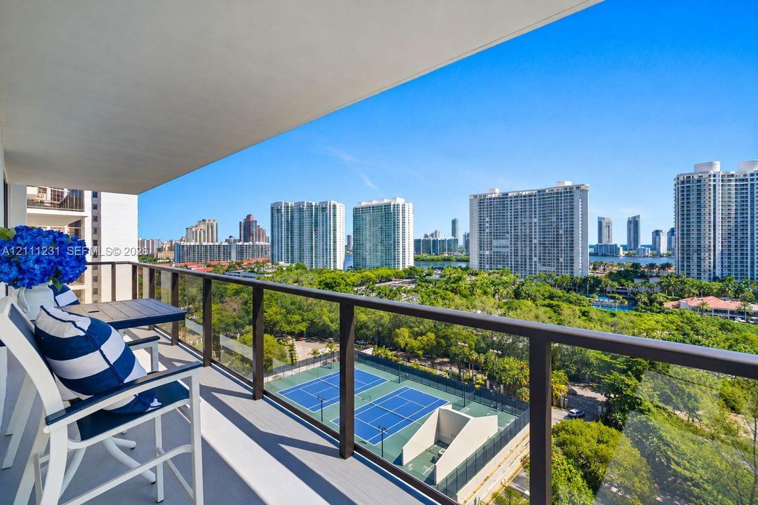 Covered private balcony featuring a glass and dark metal railing overlooking dual tennis courts and surrounding urban landscape