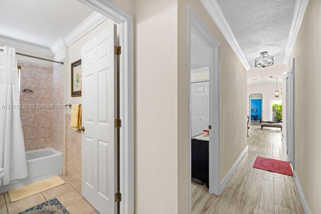 Hallway featuring wood-finish flooring, crown molding, and a multi-arm flush-mount light fixture, with a tiled bathroom including a tub-shower combination