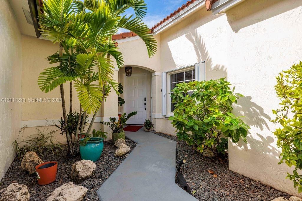 Stucco exterior entrance featuring a concrete walkway, arched entryway, and mature landscaping with rock bed mulch