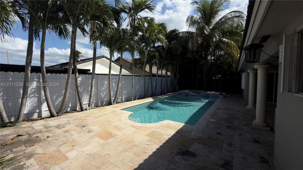 In-ground swimming pool with an integrated spa, expansive stone paver deck, white vinyl privacy fencing, and mature palm trees