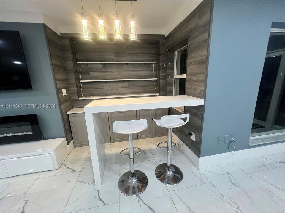 Built-in wet bar featuring a waterfall-edge countertop, grey cabinetry, integrated wood-finish wall paneling, open shelving, and multi-pendant lighting