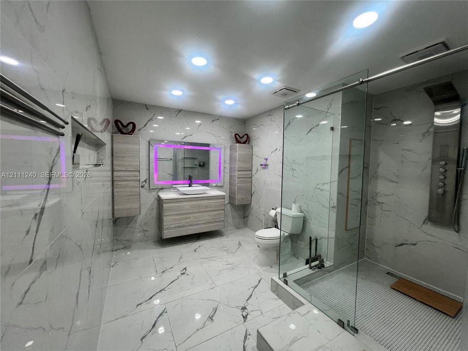 Full-height large-format wall and floor tile, wood-finish floating vanity with vessel sink, glass-enclosed shower featuring a built-in rainfall panel, recessed ceiling lighting, and wall-mounted storage cabinetry
