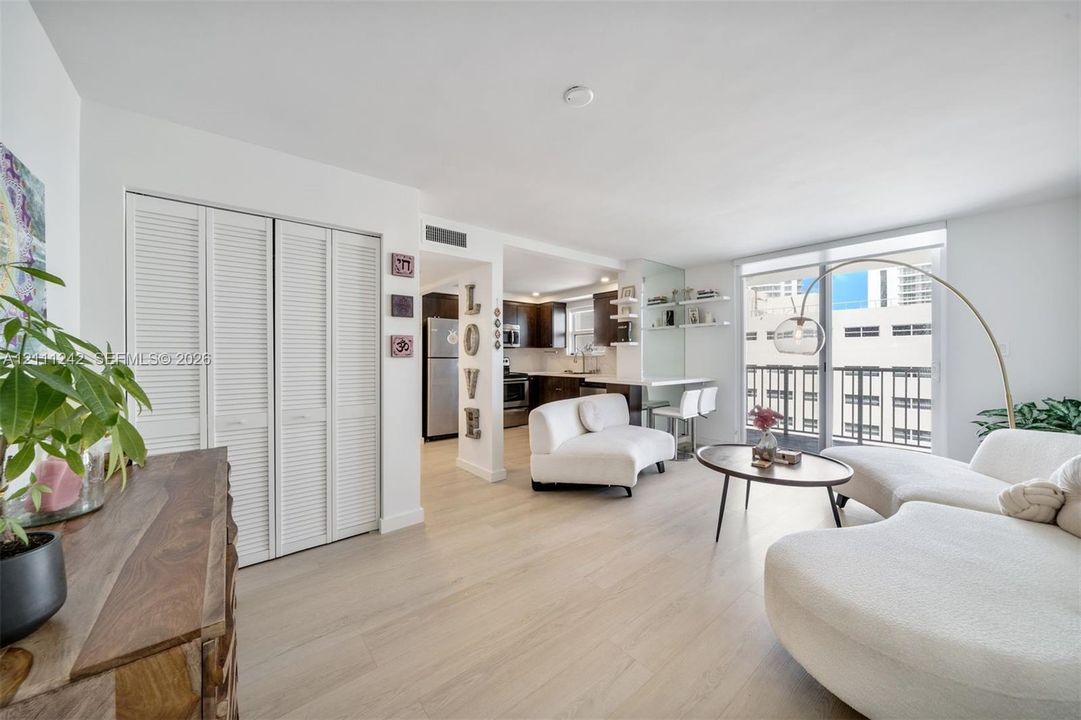 Living area featuring wood-finish flooring, floor-to-ceiling glass door, white louvered closet doors, and open-concept kitchen with dark cabinetry and built-in breakfast bar