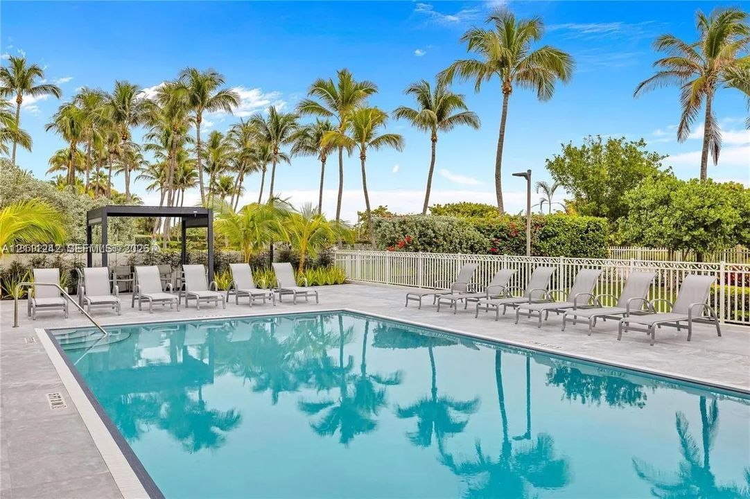 In-ground swimming pool featuring tile coping, concrete pool deck, integrated pergola, and metal perimeter fencing surrounded by mature palm trees
