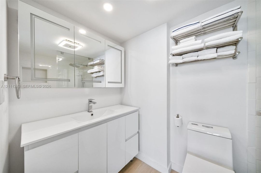 Glossy white vanity cabinet with integrated countertop, inset sink, polished chrome faucet, multi-panel medicine cabinet, and wall-mounted stainless steel shelving