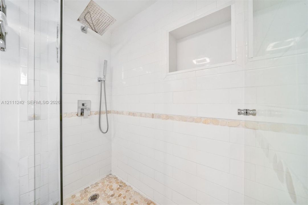 Walk-in shower featuring white subway wall tile, a built-in recessed storage niche, a river stone floor, a rainfall showerhead, and a handheld sprayer