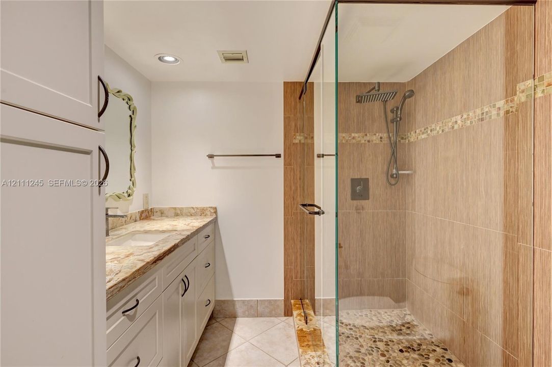 Bathroom featuring a white vanity with stone countertop, glass-enclosed shower with tiled walls, rainfall showerhead, and pebble-style shower flooring