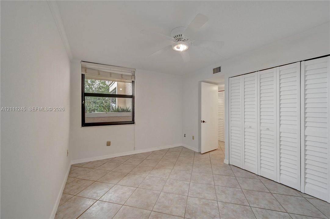 Neutral-toned tile flooring, white shutter-style closet doors, single-window illumination, and a ceiling-mounted fan with integrated lighting