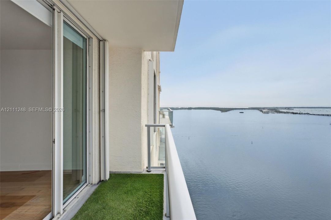Covered balcony featuring wood-finish flooring, artificial turf, and a metal railing, overlooking the open water