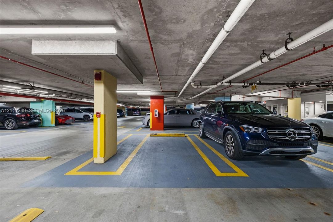 Covered parking garage featuring painted concrete flooring with designated stall markings, structural support pillars, and exposed utility piping