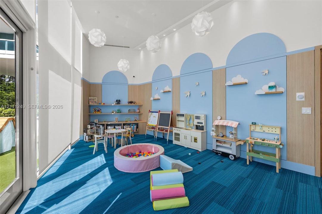 High-ceiling room featuring blue and wood-finish wall paneling with arched detailing, geometric pendant lighting, floor-to-ceiling glass paneling, and wall-mounted shelving