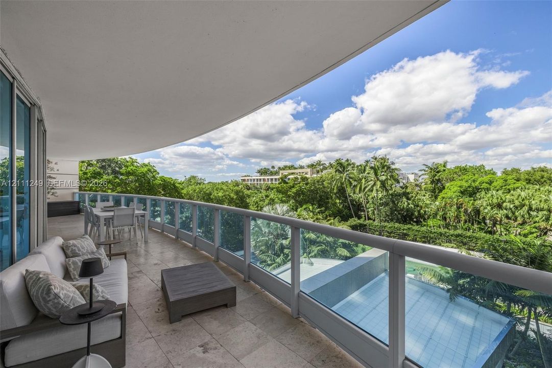 Expansive covered balcony featuring stone flooring, a glass-panel railing, and direct exposure to mature palm trees and landscaping