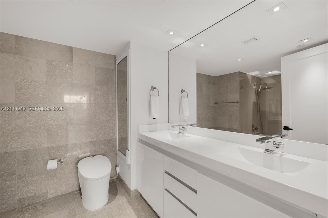 White vanity with an integrated sink and countertop, large wall-spanning mirror, recessed ceiling lighting, and floor-to-ceiling stone-tiled walls