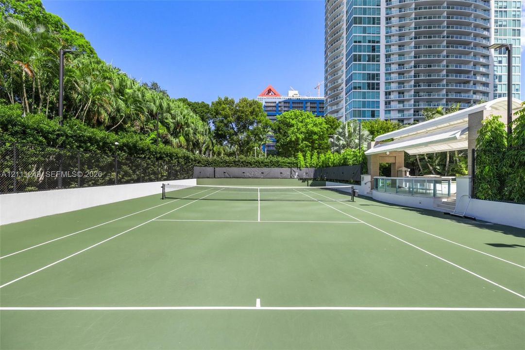 Outdoor tennis court featuring a hard surface, perimeter fencing, and an integrated pavilion with a covered seating area