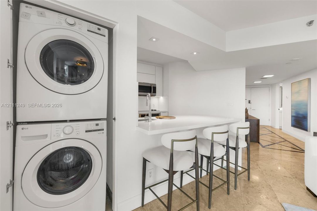 Stacked laundry unit featuring a white front-load washer and dryer, stone-surface breakfast bar with integrated sink, and stone tile flooring