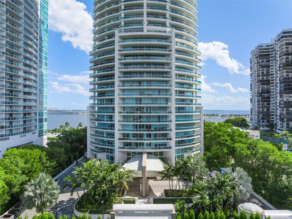 High-rise residential tower featuring a glass and concrete facade, wrap-around balconies with glass railings, a covered porte-cochère entry, and landscaped grounds