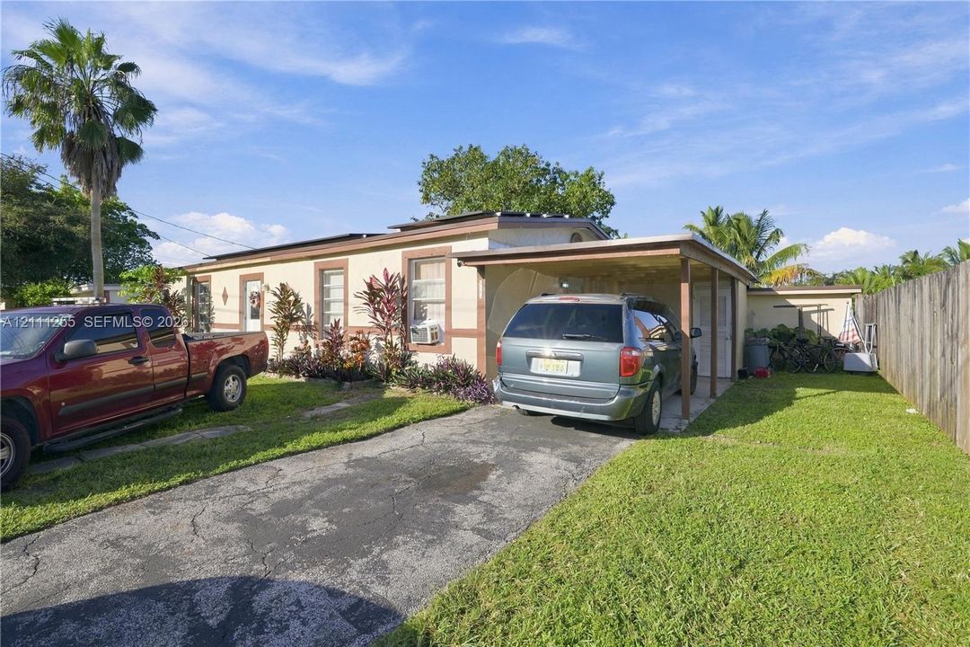 Single-story exterior featuring a paved driveway, attached carport, perimeter wood fencing, and mature palm landscaping
