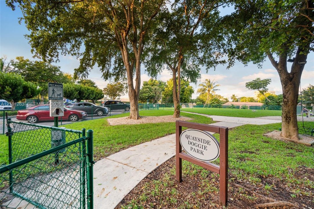 Enclosed recreation area featuring green space, concrete pathways, mature trees, and chain-link fencing