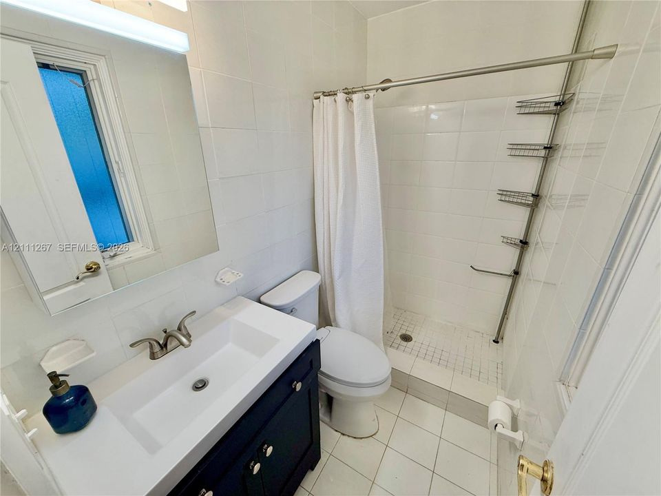 Bathroom featuring a dark-toned vanity with a rectangular sink, brushed metal faucet, white tile wall surfacing, walk-in shower with tiled surround, and integrated cabinetry