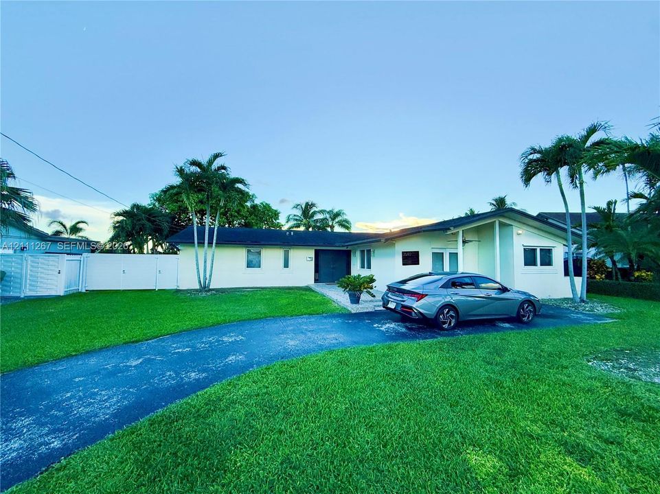 Single-story exterior featuring a light-toned facade, a curved asphalt driveway, mature palm trees, and privacy fencing