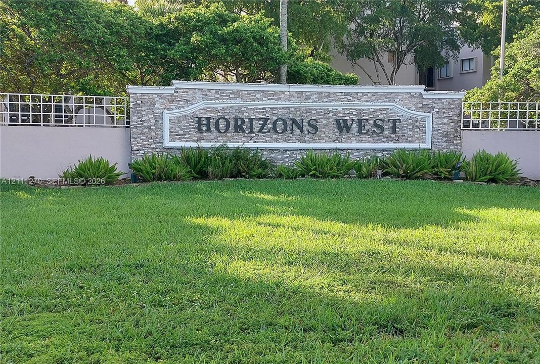 Monolithic stone-veneer community signage featuring decorative trim, bordered by a manicured lawn, established landscaping, and perimeter fencing