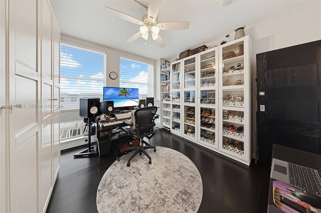 Room featuring dark-stained wood-finish flooring, floor-to-ceiling built-in white cabinetry with glass-front doors, tall windows with horizontal blinds, and a multi-blade ceiling fan with integrated lighting