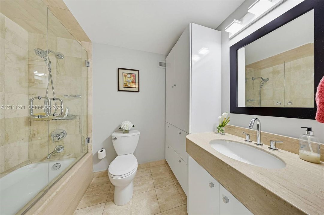 Bathroom featuring stone tile tub surround, frameless glass shower door, floor-to-ceiling built-in storage cabinetry, and vanity with an integrated stone countertop and oval undermount sink