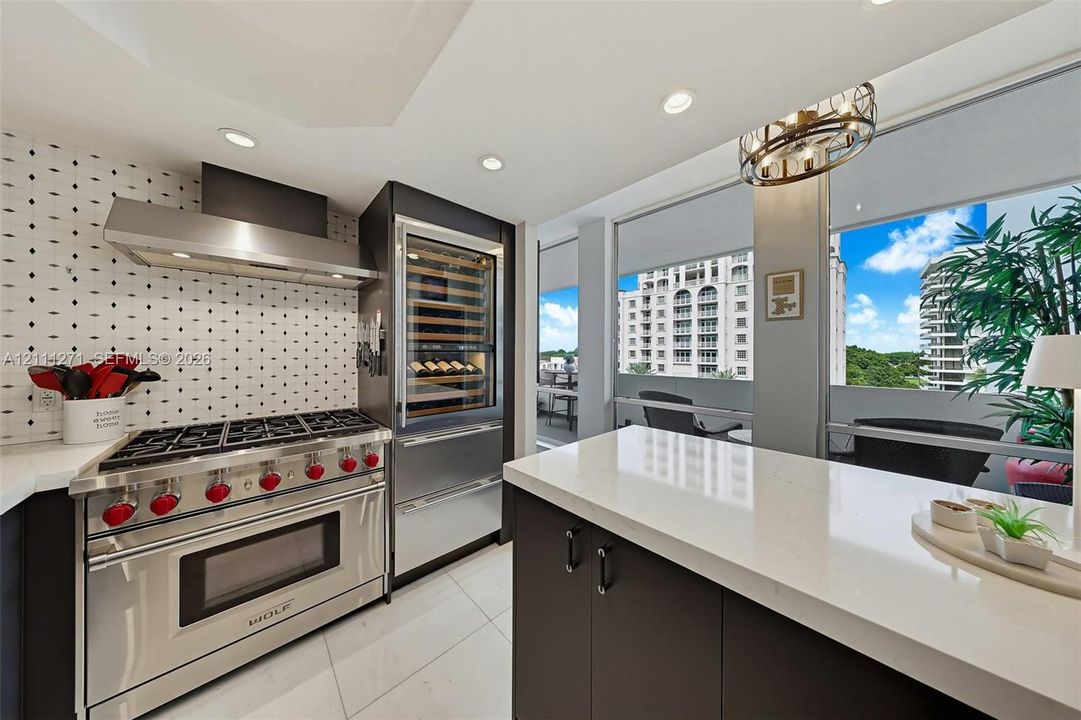 Kitchen featuring a stainless steel Wolf gas range, integrated wine refrigeration, stone-surfaced island, patterned tile backsplash, and floor-to-ceiling windows