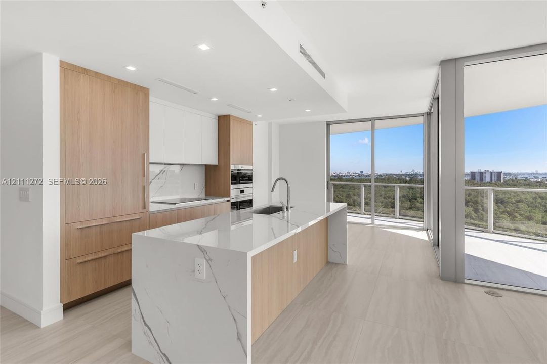 Kitchen featuring a waterfall island with a stone-finish countertop, integrated wood-finish cabinetry, built-in double wall ovens, and floor-to-ceiling glass doors opening to an exterior balcony