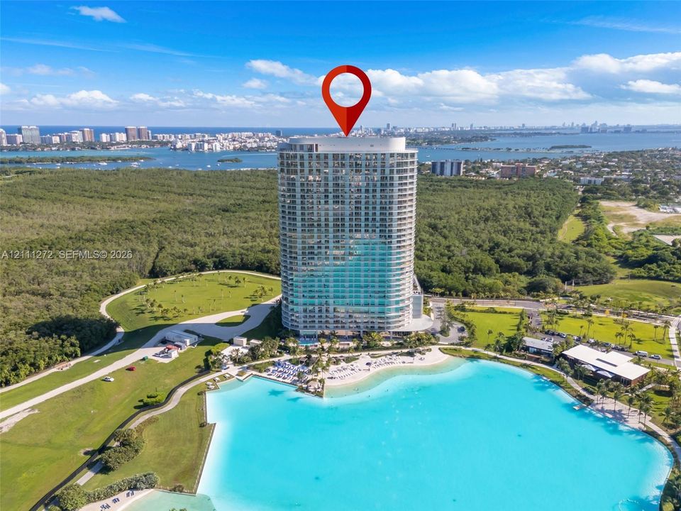 High-rise residential tower featuring a glass facade, situated on a waterfront lot with an expansive turquoise lagoon, surrounded by established greenery and winding paved pathways