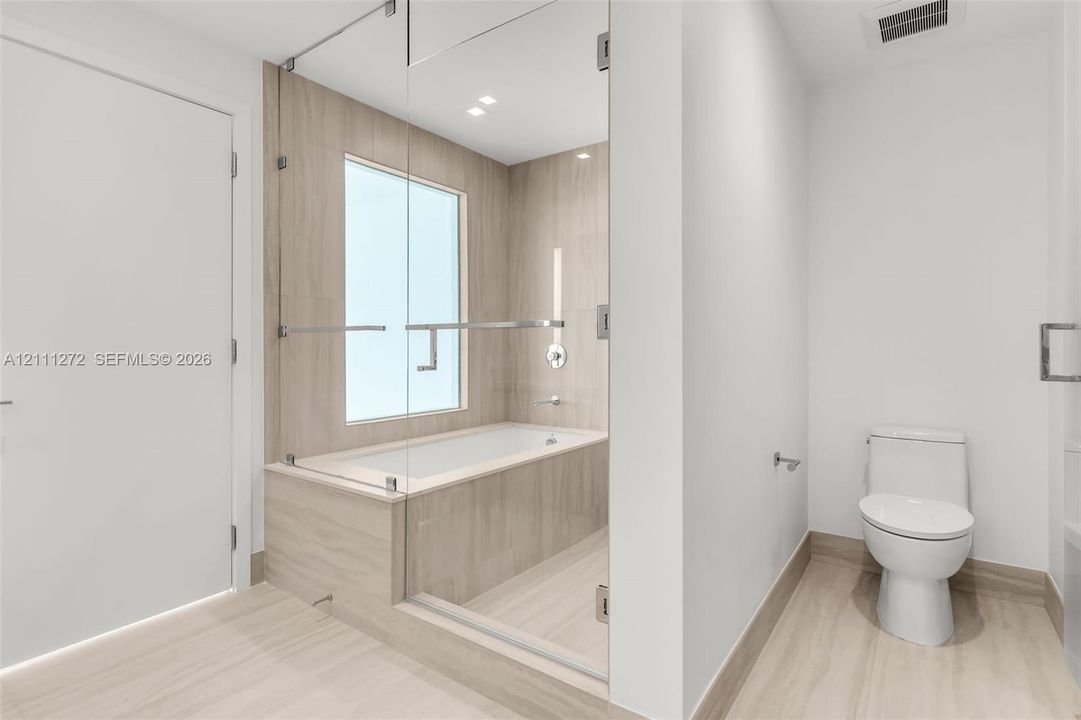 Bathroom featuring a frameless glass-enclosed tub with floor-to-ceiling tile surround, neutral-toned large-format tile flooring, and a white porcelain commode