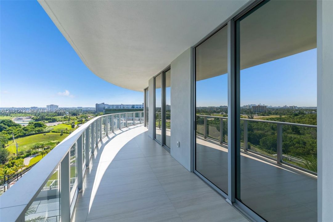 Expansive covered balcony featuring large-format tile flooring, a curved architectural design, and glass panel railing with metal supports, complemented by floor-to-ceiling sliding glass doors