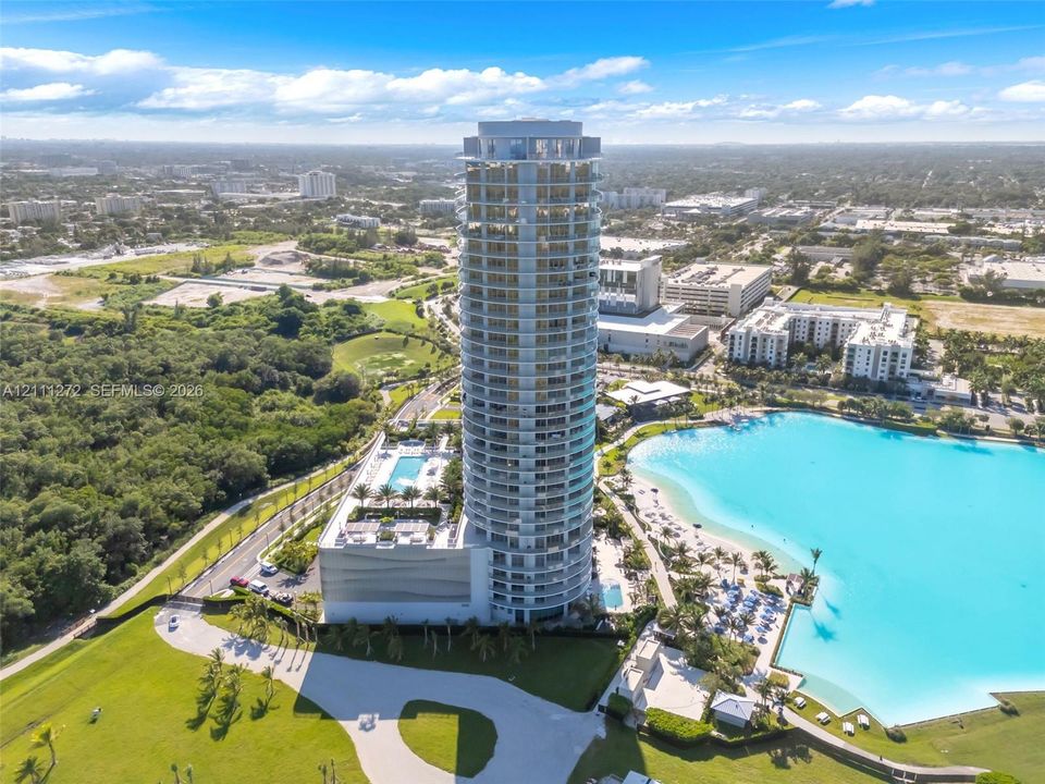High-rise residential tower featuring a cylindrical glass facade, surrounding private balconies, a ground-level swimming pool, and immediate proximity to a turquoise lagoon