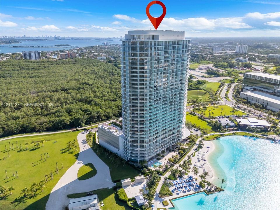 High-rise glass facade building featuring wrap-around balconies, a ground-level swimming pool, and immediate access to a man-made lagoon with a sand shoreline, set amidst extensive landscaped grounds