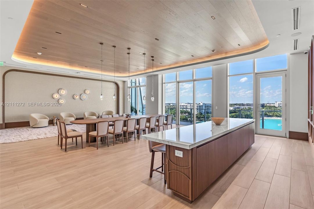 Expansive space featuring a curved wood-slat ceiling with recessed perimeter lighting, floor-to-ceiling windows, wood-finish tile flooring, and a large central island with a stone-top surface and wood cabinetry