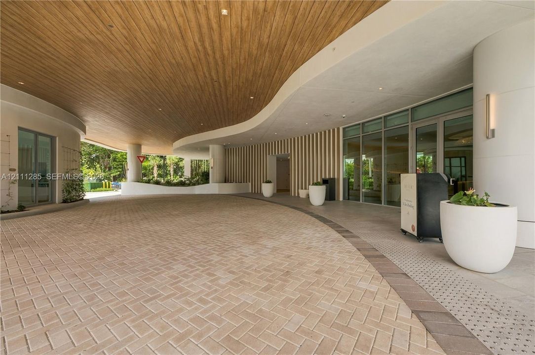 Covered porte-cochère featuring a curved wood-panel ceiling, herringbone-pattern paver drive, vertical slat wall detailing, and expansive glass entry doors