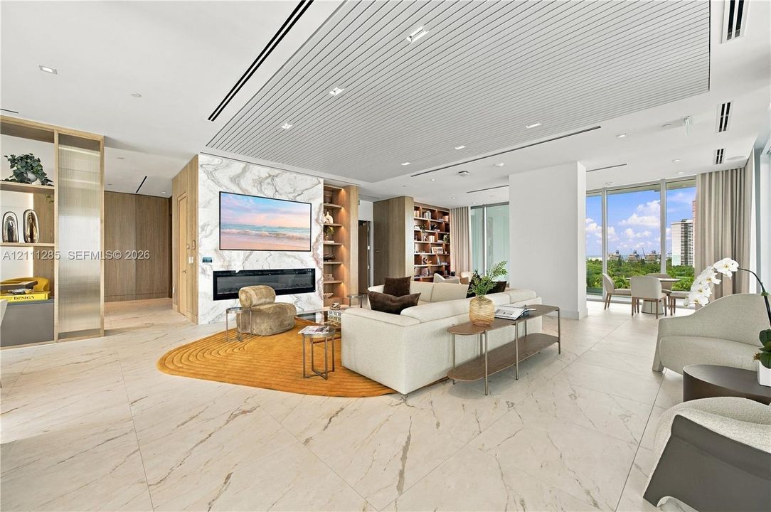 Open-concept interior featuring large-format tile flooring, a floor-to-ceiling stone-clad fireplace wall, custom built-in shelving, a tiered slatted ceiling with recessed lighting, and floor-to-ceiling windows