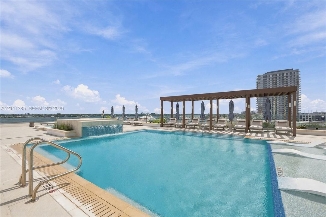 Rooftop swimming pool featuring a waterfall edge, integrated shallow sun shelf, metal handrails, and a covered pergola