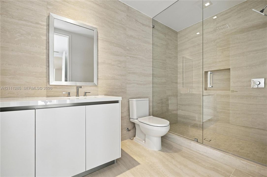 Wall-to-wall stone tile surfacing, walk-in glass-enclosed shower with a recessed shelf, white vanity with a rectangular sink, wall-mounted mirror, and a two-piece toilet