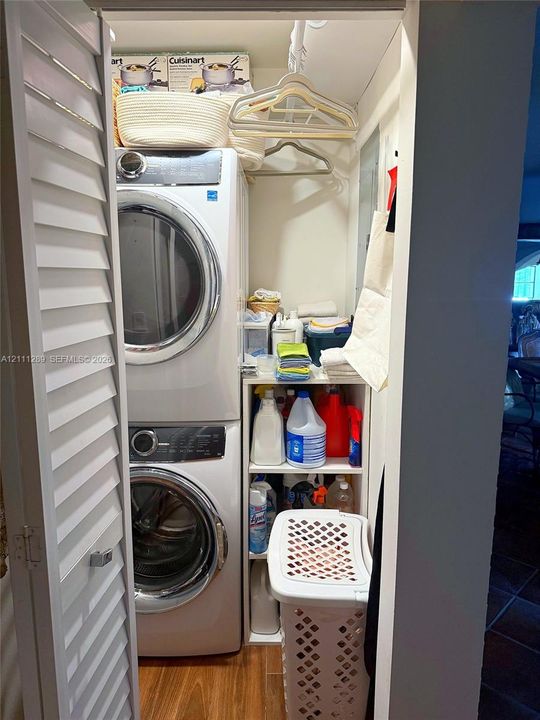 Laundry closet featuring stacked front-load washer and dryer, louvered bi-fold doors, wood-finish flooring, and built-in vertical shelving